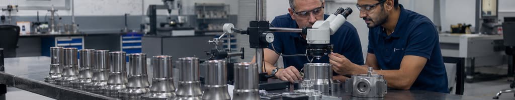 Quality engineer inspecting a finished metal component under magnification after production, recording findings during a laboratory investigation.